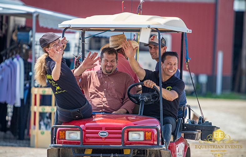 Three people ride in a red utility cart, smiling and waving under a white canopy.