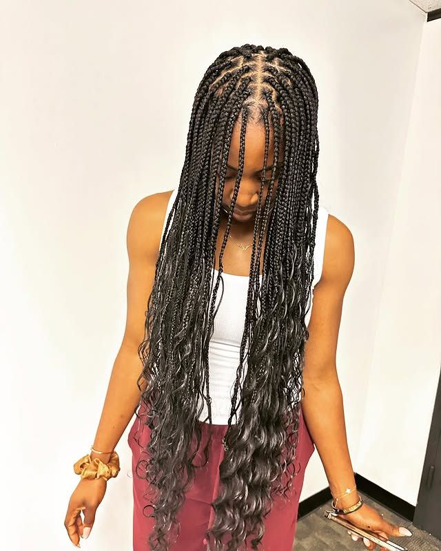Woman with long braided hair looking down, wearing a white tank top and burgundy pants indoors.