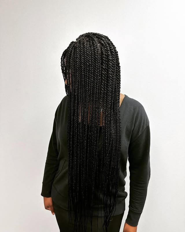 Person standing with long black braided hair cascading down the back, wearing a dark shirt against a white wall.