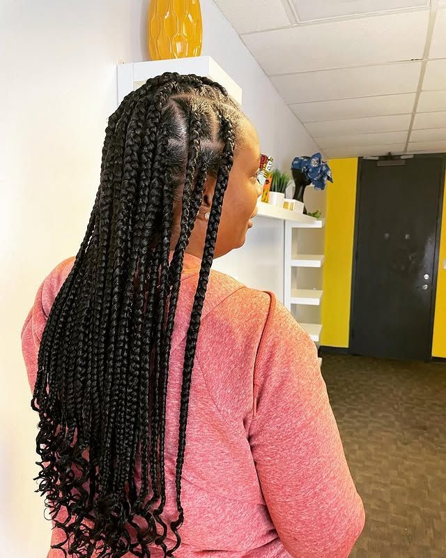Person with long braids in a pink sweater stands indoors near a white shelf and yellow wall.