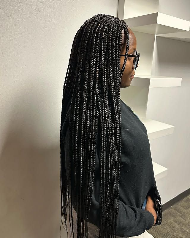 Person with long braided hair and glasses in a black outfit, standing in profile indoors.
