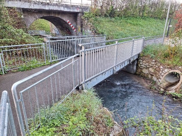 Metallene Fußgängerbrücke über einen schmalen Bach neben einer Steinbogenbrücke und einem grasbewachsenen Ufer