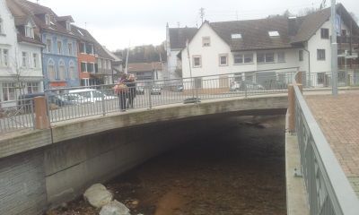 Fußgängerbrücke über einen schmalen Fluss in einer Kleinstadt, im Hintergrund Gebäude und einige Menschen.