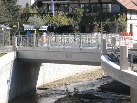 Eine Fußgängerbrücke aus Beton führt über einen schmalen Kanal neben Gebäuden und Bäumen.
