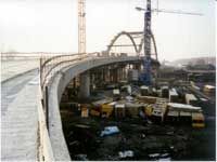Gebogene, erhöhte Brücke im Bau, Kräne und schweres Baugerät neben einem Flussufer