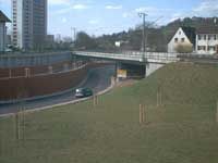 Kurvenreiche Straße unter einer Autobahnbrücke neben einem grasbewachsenen Hang, im Hintergrund ein Auto und Häuser.