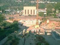 Große Baustelle mit Baggern und Kränen neben einem hohen beigefarbenen Gebäude, von oben betrachtet.