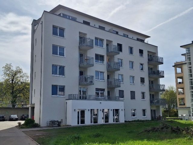 Fünfstöckiges weißes Wohnhaus mit Balkonen und grasbewachsenem Vordergrund unter bewölktem Himmel