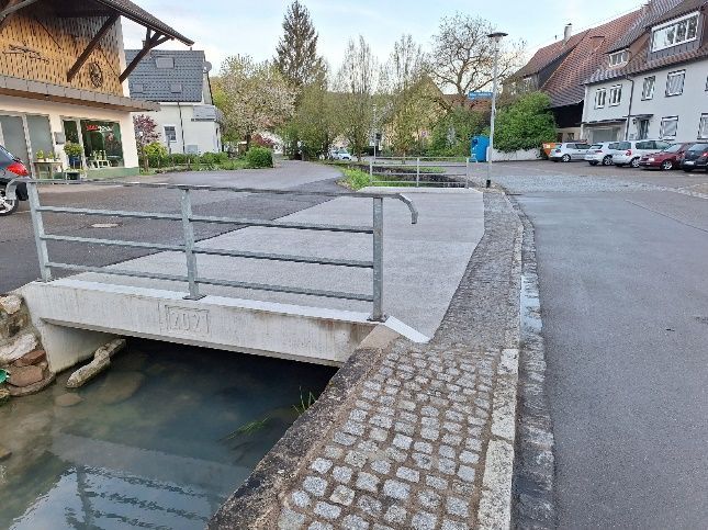 Rampenförmige Gehwegbrücke über einen Entwässerungskanal neben einer Kopfsteinpflasterstraße in einem Dorfviertel