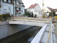 Kleine Betonbrücke über einen Kanal in einem Wohngebiet, in der Nähe von Häusern und Geländern.