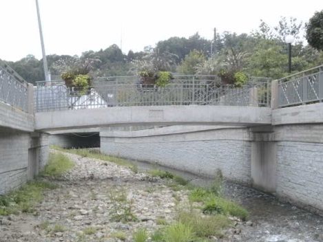 Betonkanal unter einer Straßenbrücke mit felsigem, trockenem Bachbett und eingezäunten Seiten, darüber Bäume