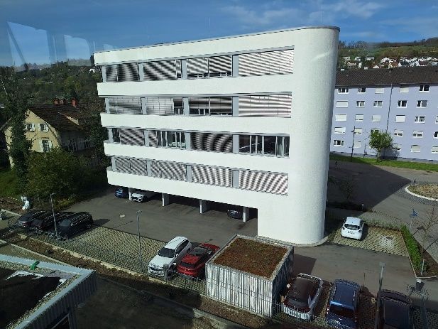 Modernes weißes Wohnhaus mit abgerundeten Ecken und gestreiften Balkonen, über den geparkten Autos zu sehen.