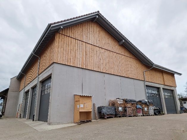 Große graue Lagerhalle mit Holzverkleidung und Müllcontainern im Freien unter einem bedeckten Himmel