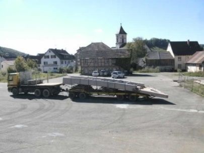 Ein Pritschenwagen transportiert eine lange Ladung auf einem Dorfplatz mit Häusern und einer Kirche im Hintergrund.