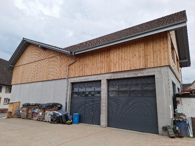 Modernes Hausäußeres mit grauen Garagentoren, hölzerner Oberfassade und Mülltonnen entlang der Einfahrt