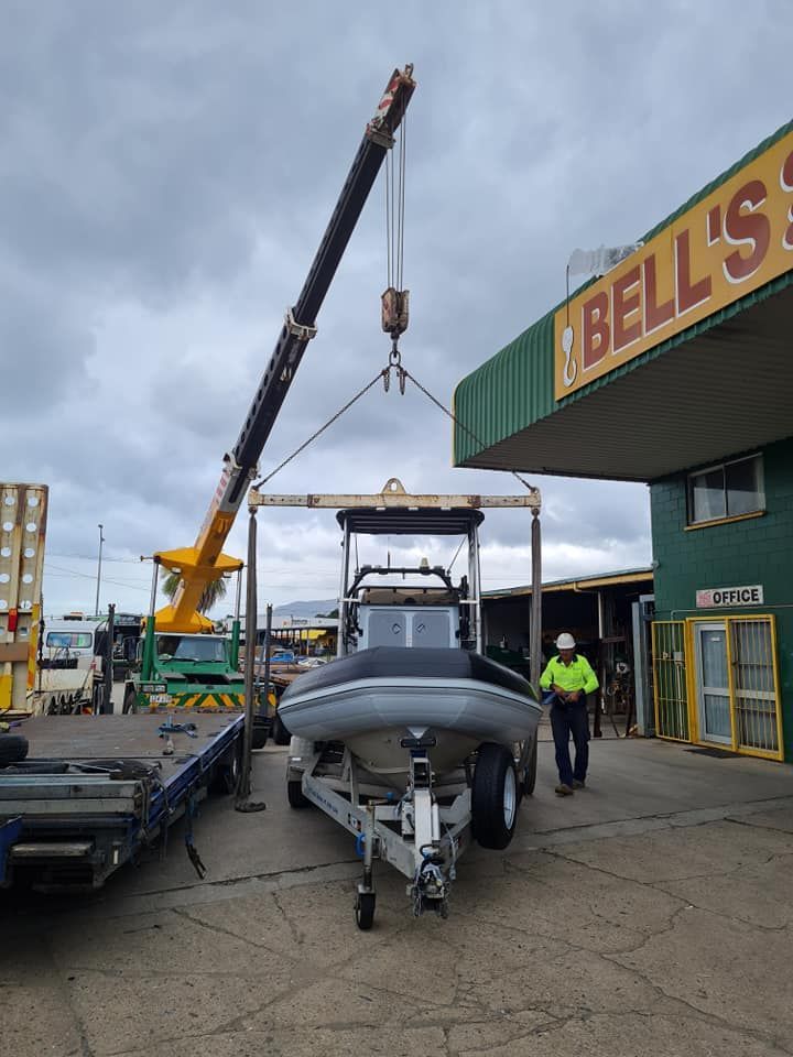 Crane lifting a gray speedboat beside a Bell’s sign — Bell's Carrying & Crane Hire Service Pty Ltd in Atherton, QLD