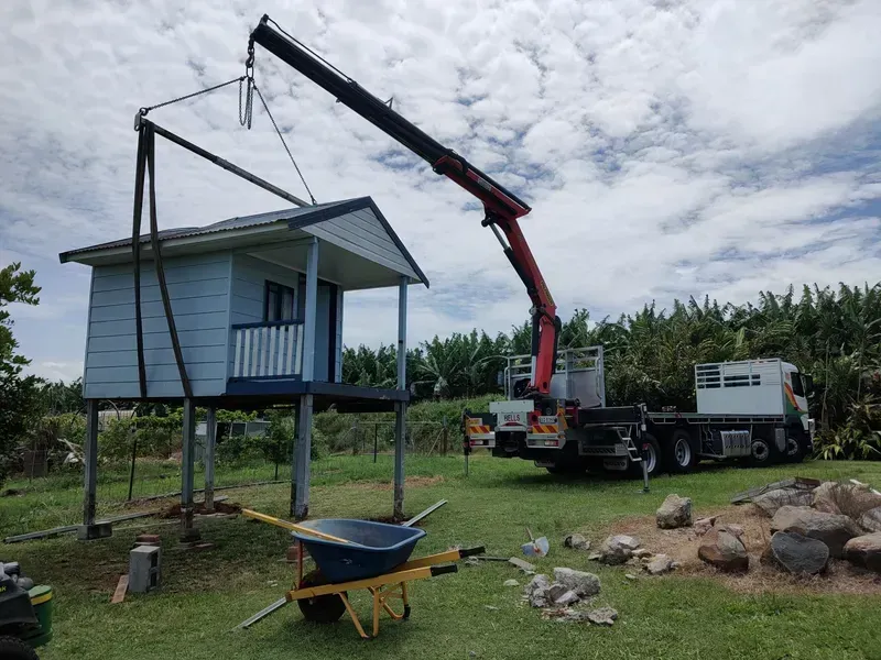 A Crane Lifting a Small, Blue House on Stilts — Bell's Carrying & Crane Hire Service Pty Ltd in Bungalow, QLD