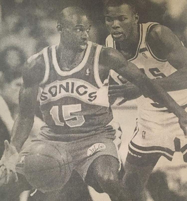 Basketball players, one in a Sonics jersey with the ball, defended by another in a jersey. Court setting.