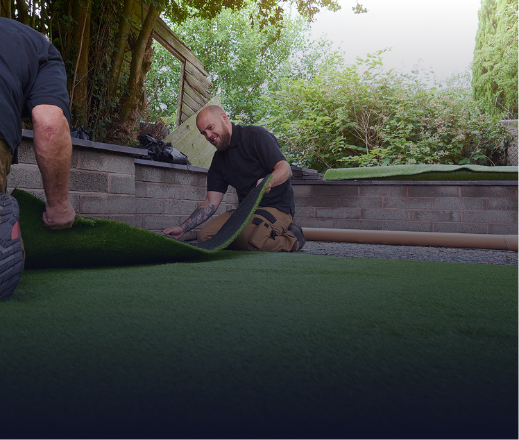 Two people installing artificial turf on a patio with a brick wall and trees in the background.