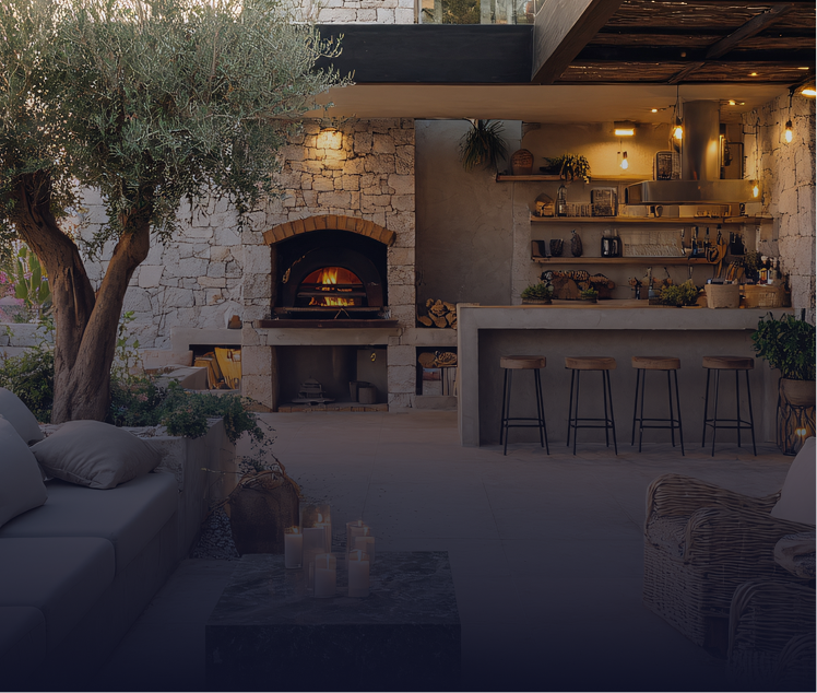 Outdoor kitchen with brick oven, bar, seating, and olive tree. Evening scene.