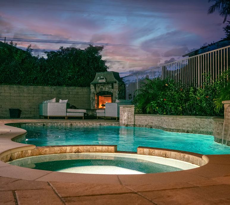 A backyard pool with a hot tub and fireplace, under a twilight sky.