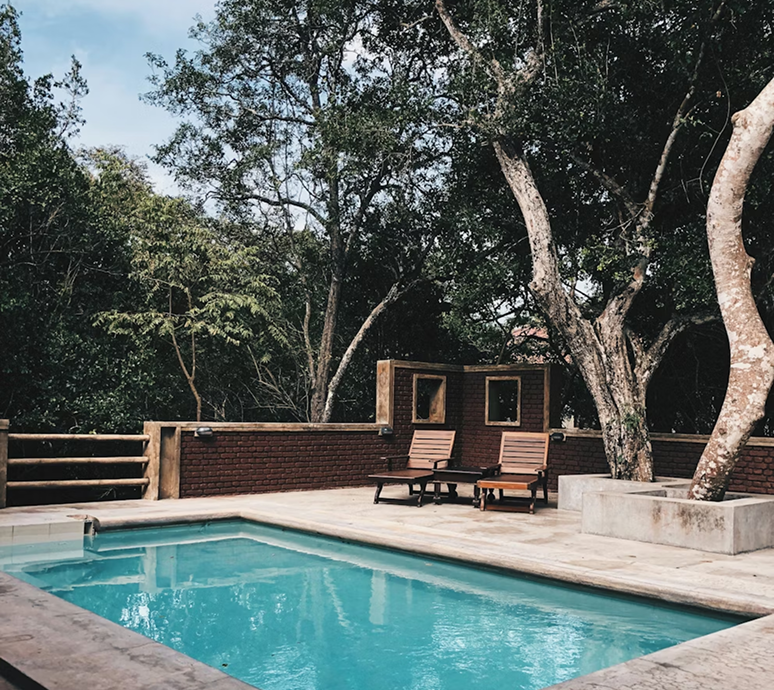 A turquoise pool surrounded by a brick wall, chairs, and trees in a natural setting.