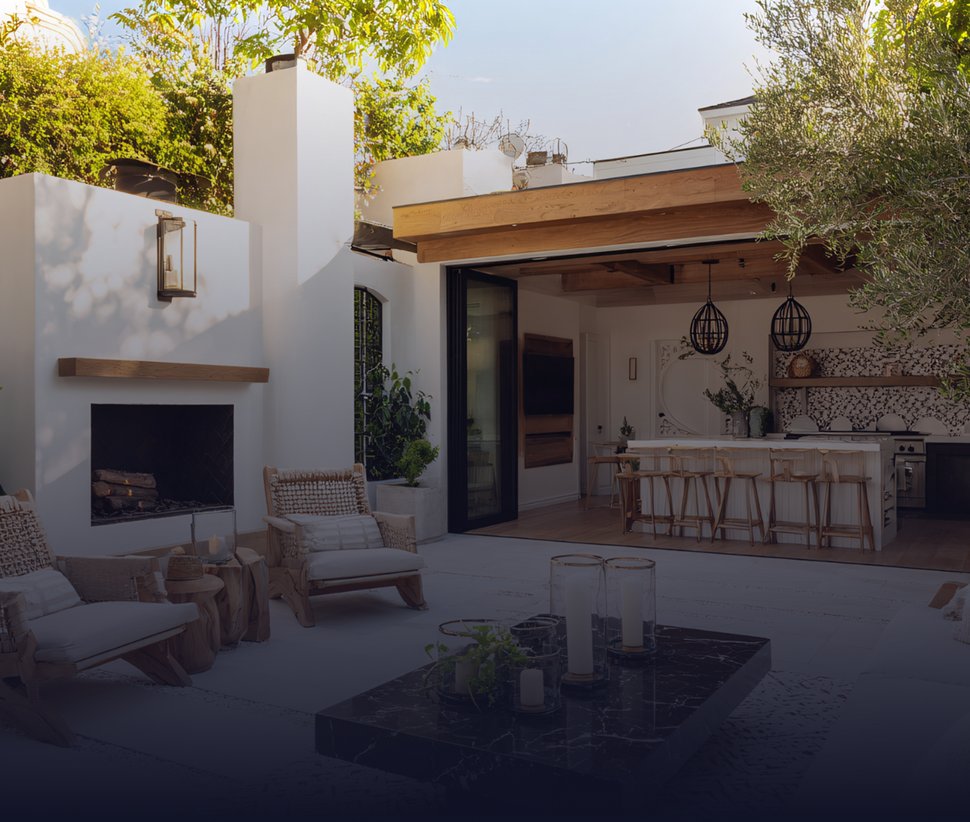 Outdoor patio with fireplace, kitchen, and seating area. White walls, wooden accents, and greenery.
