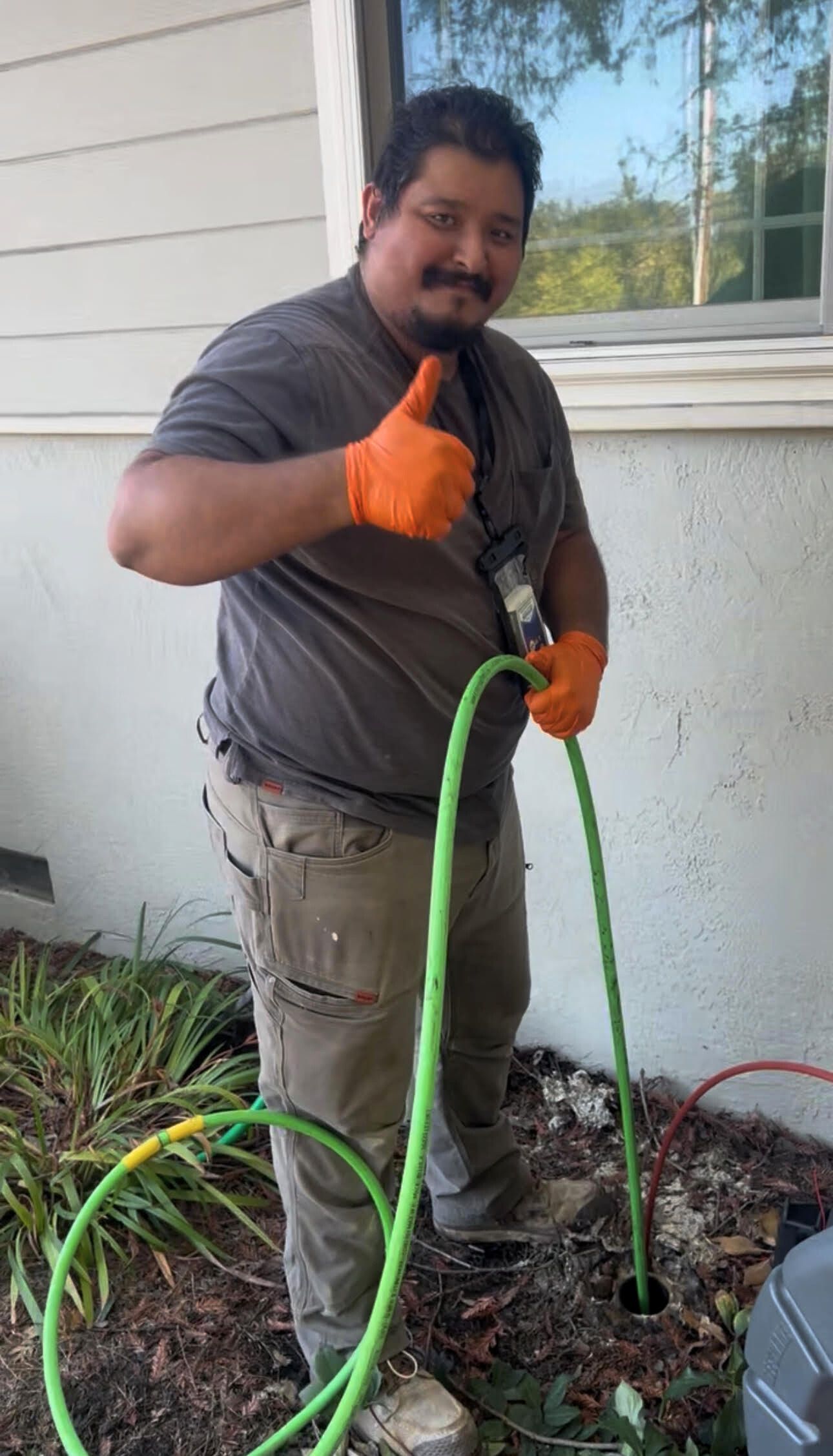 Man in work clothes giving a thumbs-up while holding green hose near the ground and building. Orange gloves.