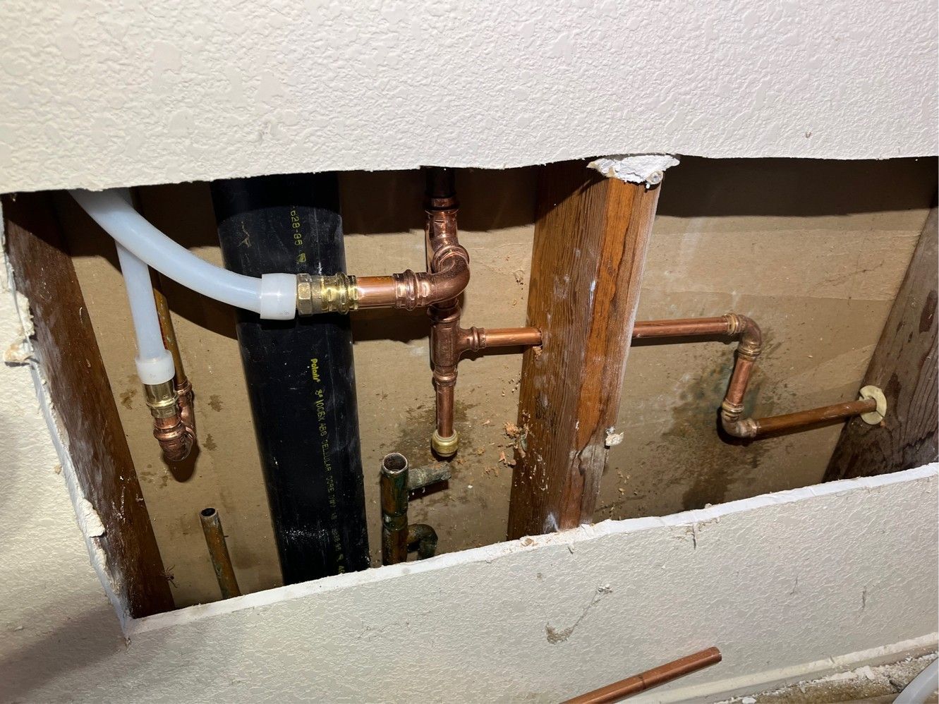 Copper pipes and fittings in a wall opening, connected to black pipe and white tubing.