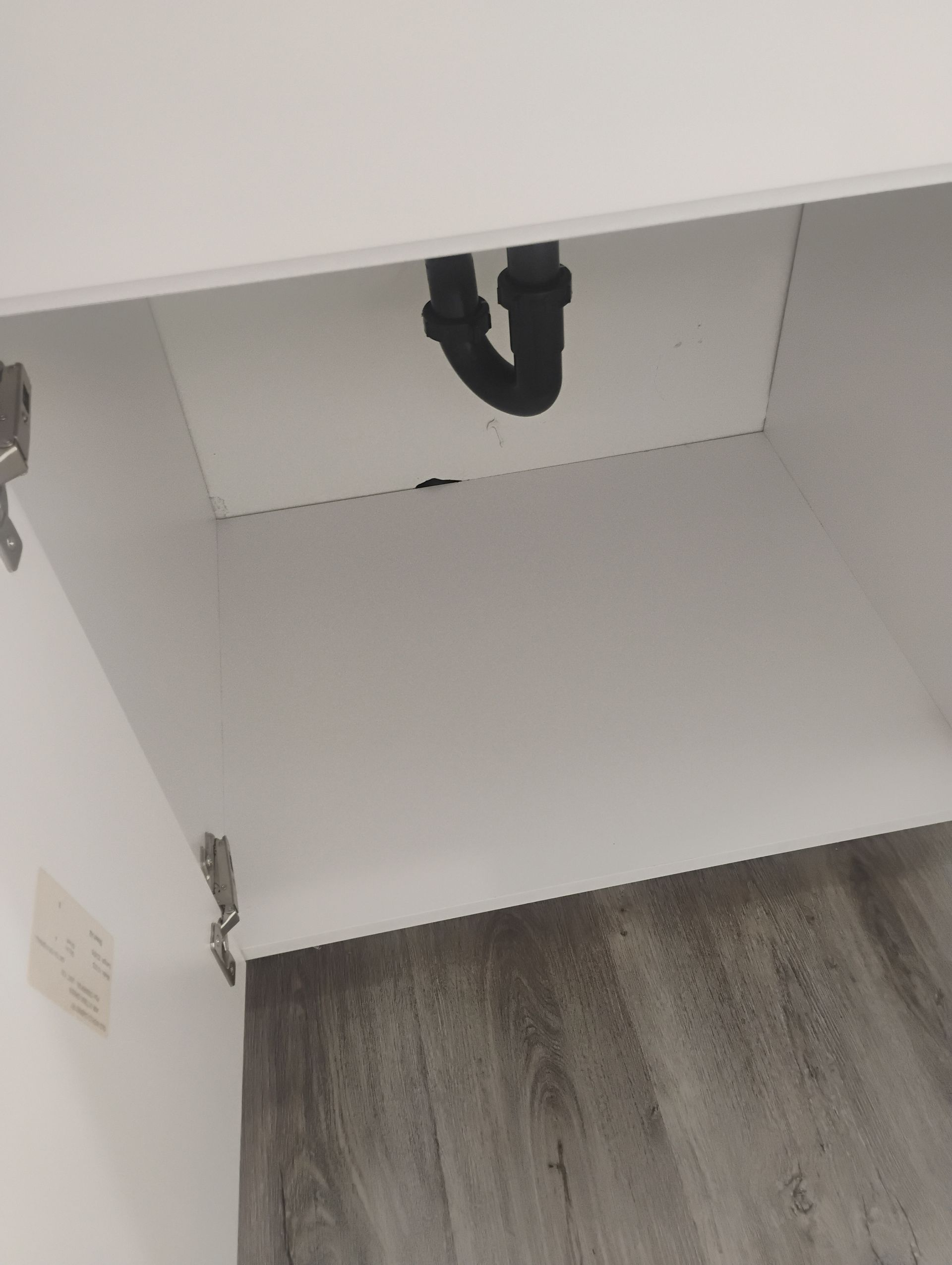 Empty cabinet under a sink with a black drain pipe and white interior, above gray wood flooring