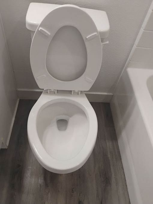 Open toilet with white seat in a small tiled bathroom beside a bathtub.