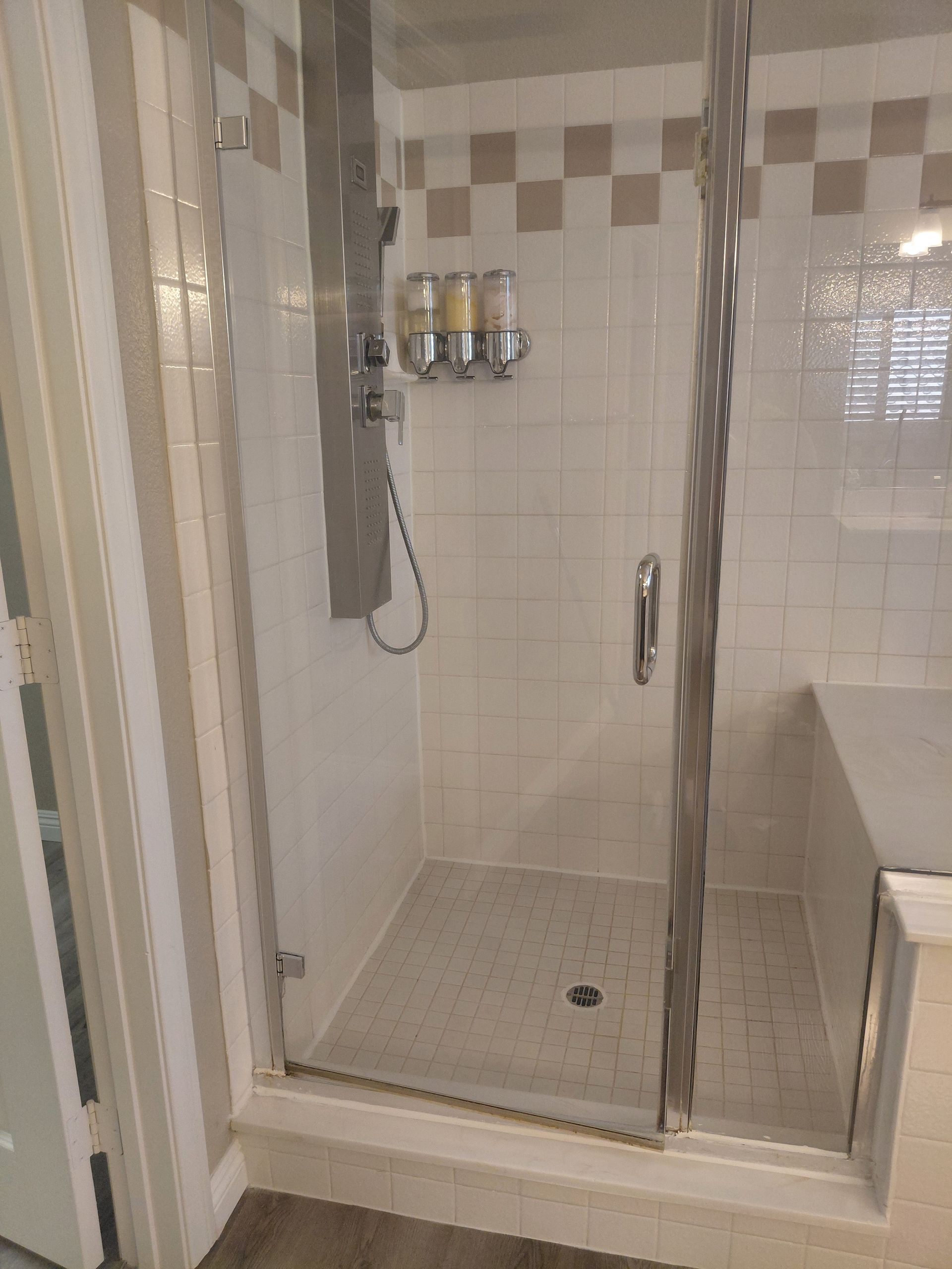 Corner shower with glass door, tiled walls, wall-mounted shower dispenser, and white bench seat.