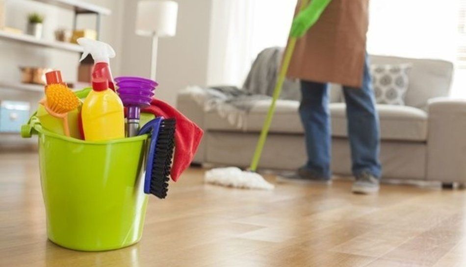 Cleaning supplies in a green bucket on a wooden floor, with a person mopping in the background.
