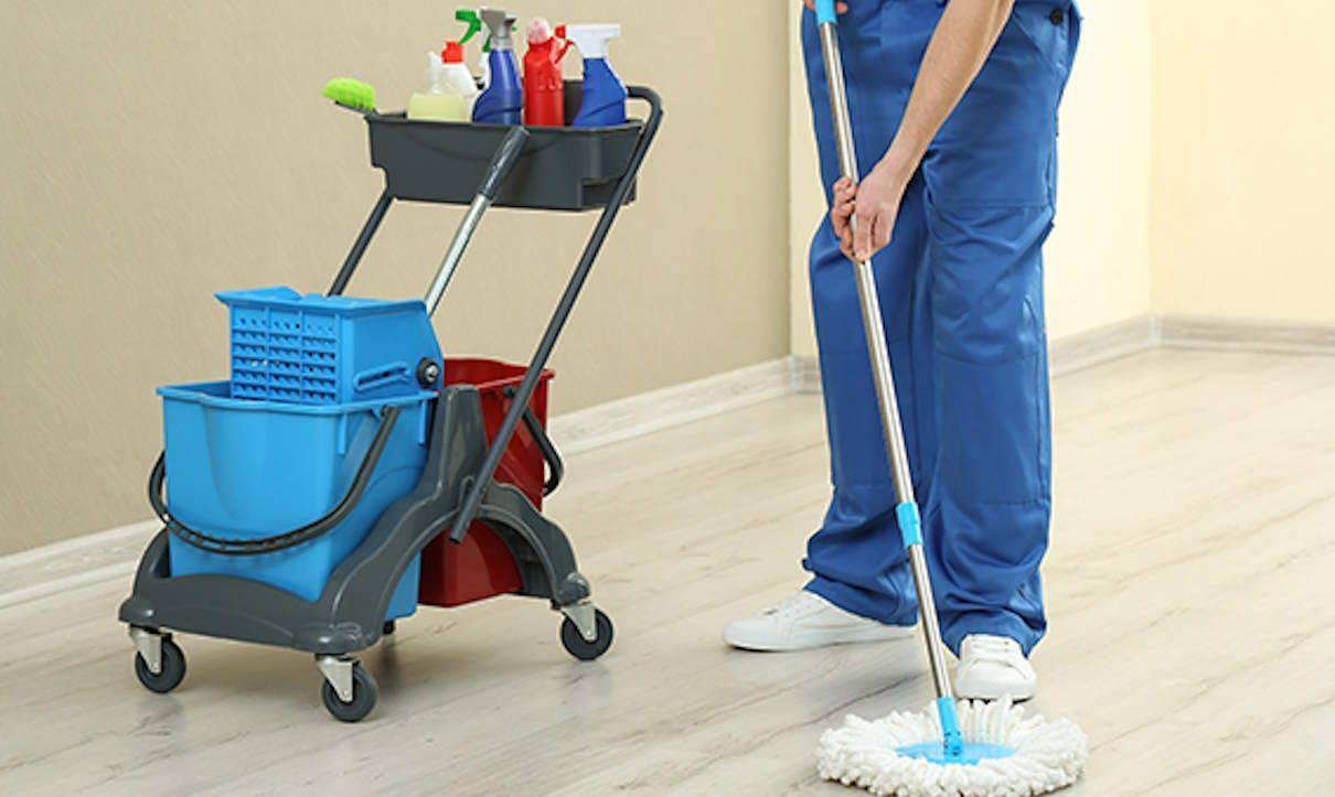 Custodian mopping a hallway floor with a blue cleaning cart nearby