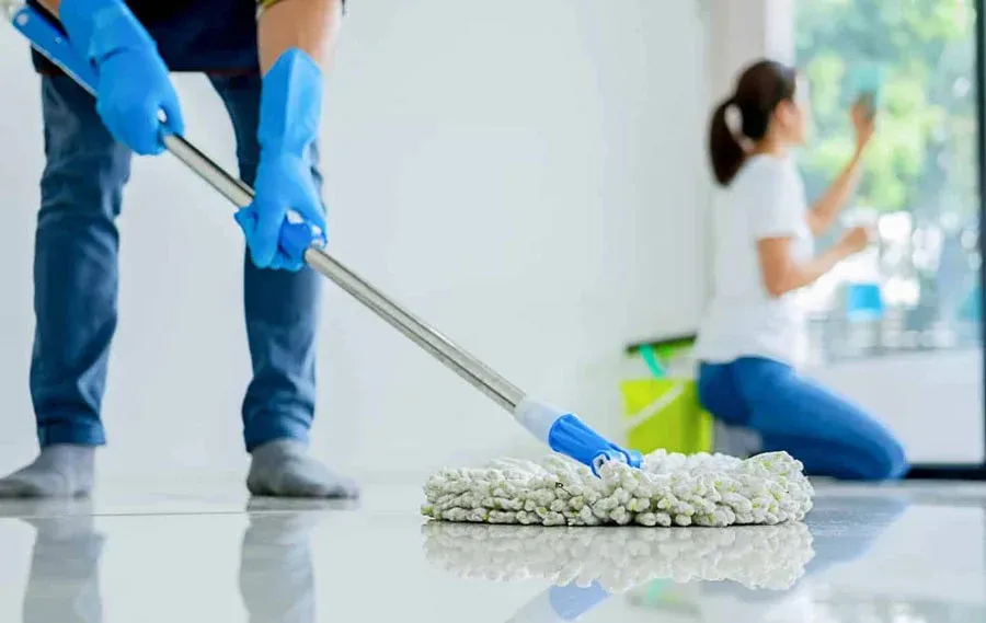 Mop cleaning a shiny floor while a person kneels in the background in a bright room