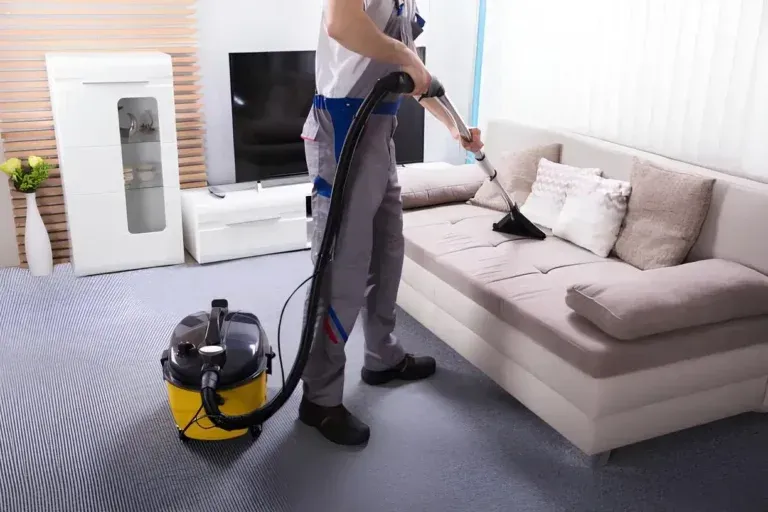 Person vacuuming a beige sofa in a bright living room with a canister vacuum cleaner