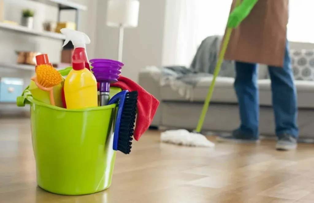 Cleaning supplies in a green bucket beside a person mopping a wooden floor in a home.