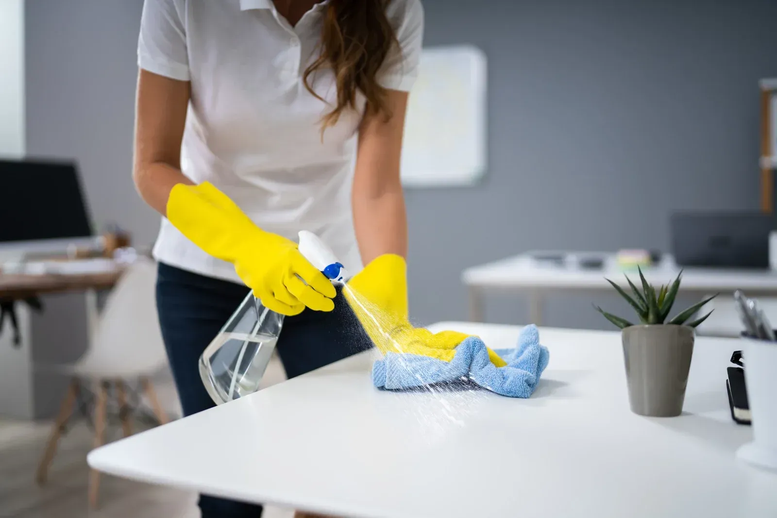 Person cleaning a white table with a spray bottle, yellow gloves, and a cloth in an office setting