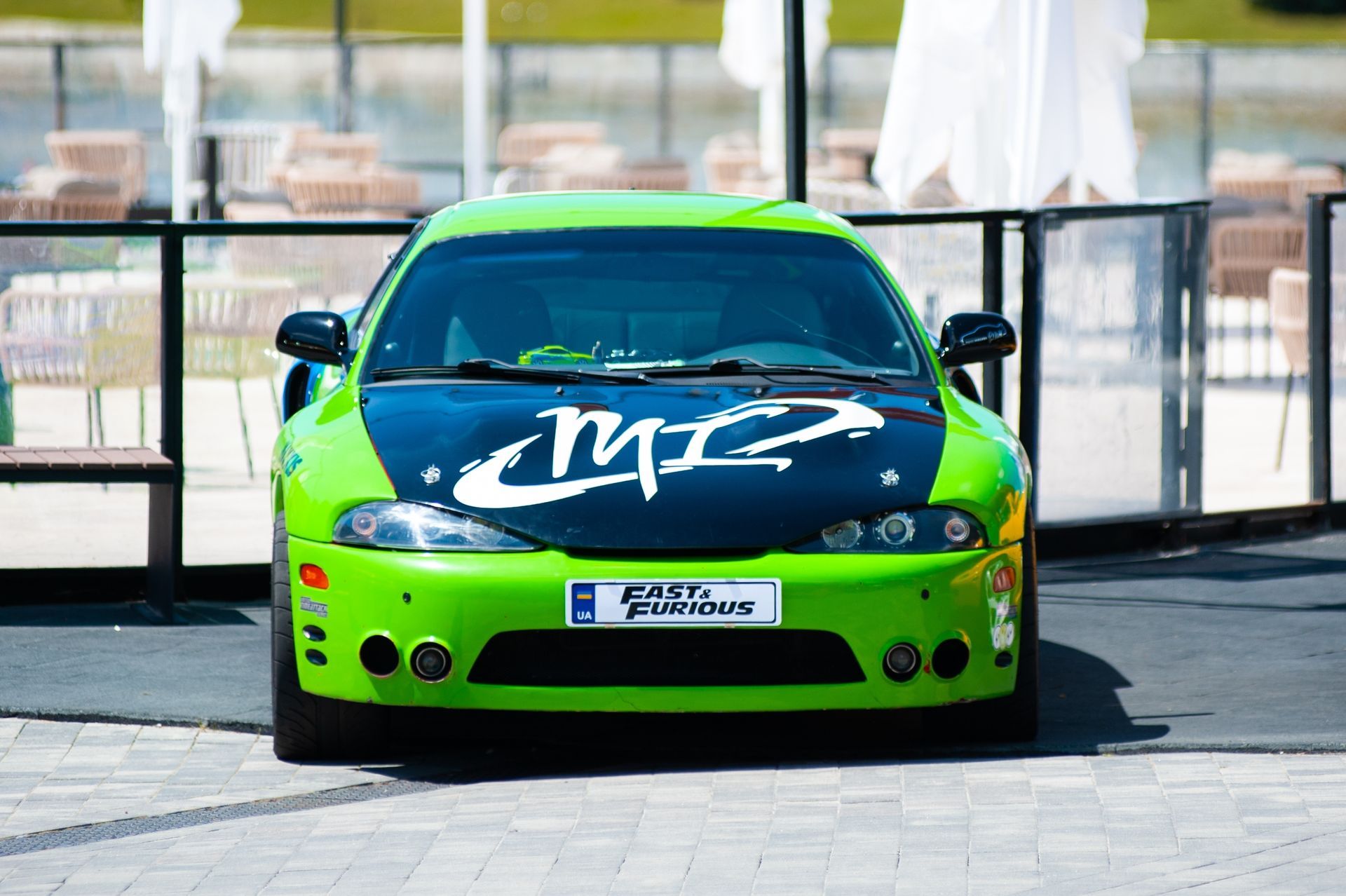 Bright green and blue sports car with “MZ” on the hood, parked outdoors behind a barrier.