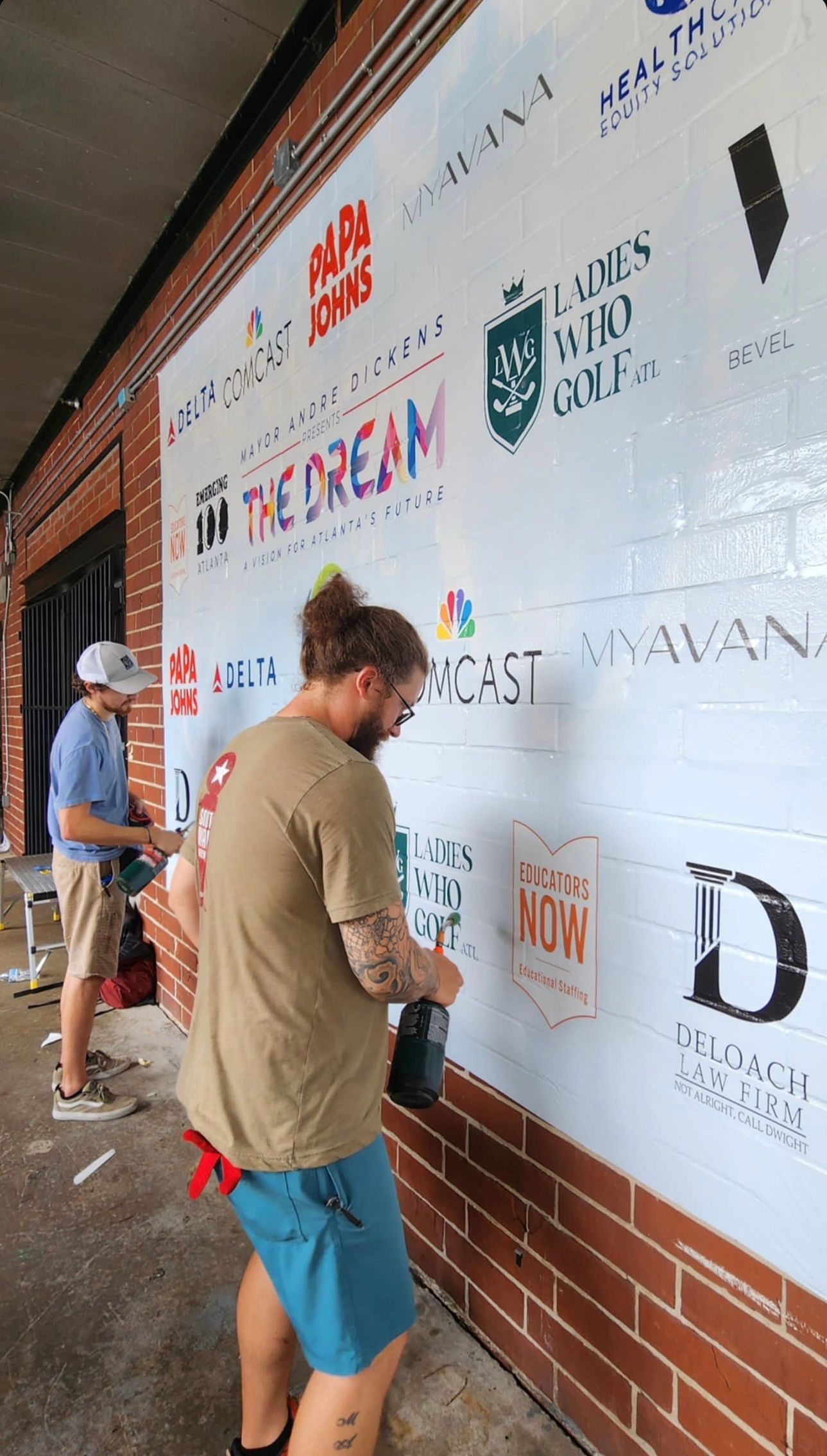 People placing logos on a white event backdrop beside a brick wall, one holding a phone under an awning.