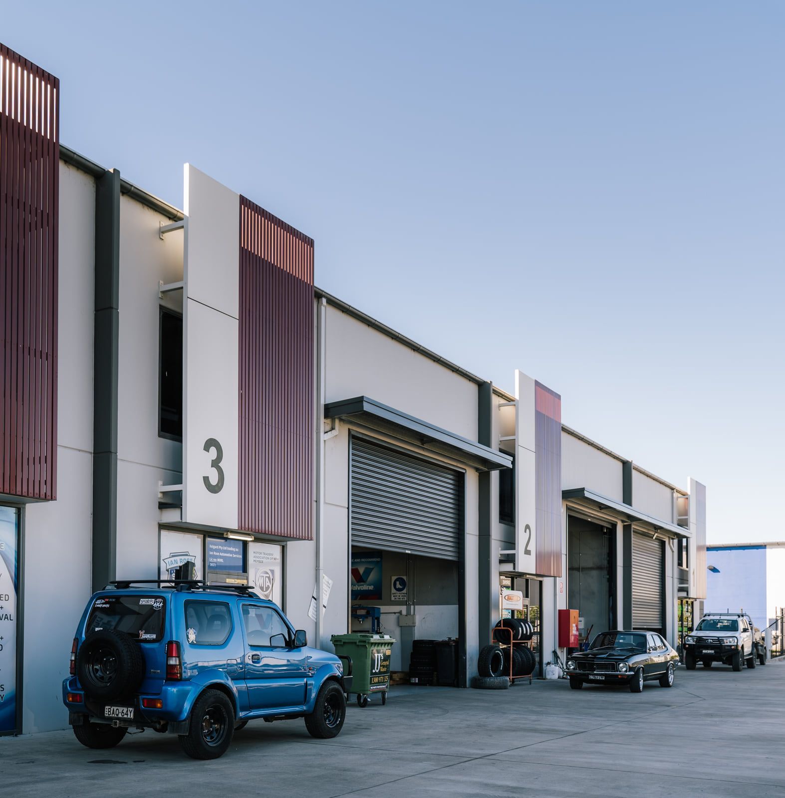 Industrial warehouse bays with parked blue and black vehicles under a clear sky — Gardiner Automotive & Performance in Rutherford, NSW