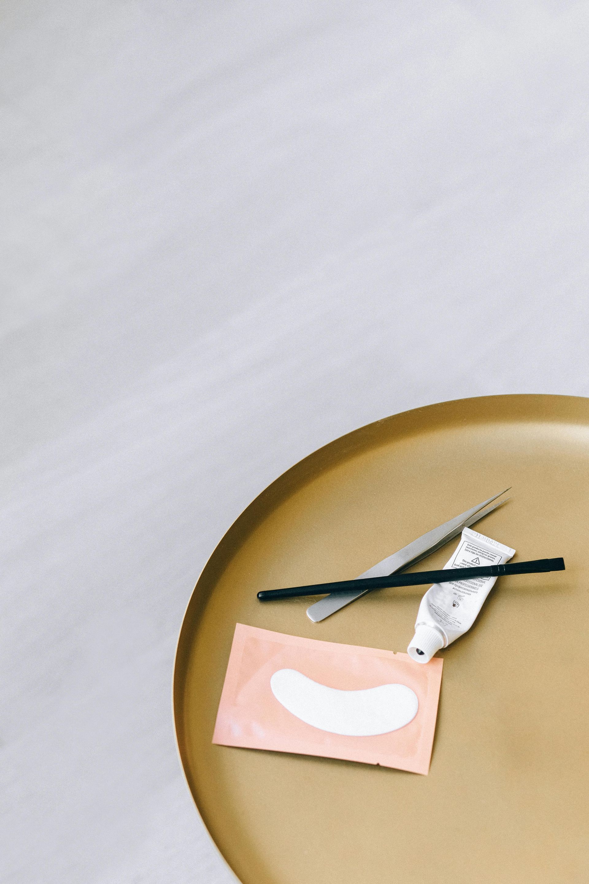 Eyelash extension tools on a gold tray: tweezers, glue tube, under-eye pads, and applicators against a white background.