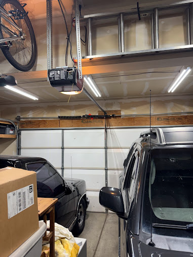 Garage interior with two parked cars, stacked boxes, a bicycle, and a ceiling-mounted garage door opener.