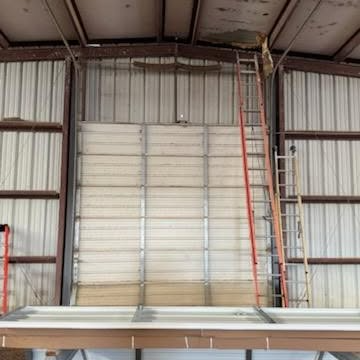Interior of a warehouse with a high corrugated wall, red ladder, and stacked materials near the ceiling