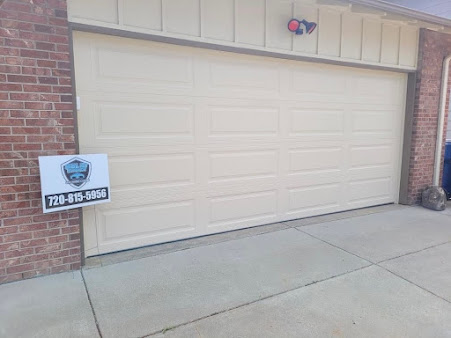 Beige garage door with a real estate sign and phone number beside a brick house