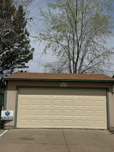 Two-car garage with beige doors and brown roof, framed by trees and a driveway