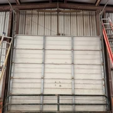 Industrial warehouse interior with a large white roll-up door centered between metal supports and a ladder on the right.