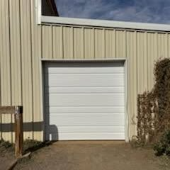 White roll-up garage door on a beige metal building with a concrete drive and bushes on the right.