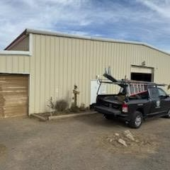 Black pickup truck parked beside a beige metal building with a garage door and ladder on the roof rack