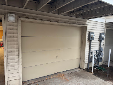 Beige garage door under a covered carport with utility meters and pipes on the right